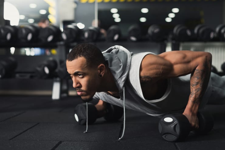 An athlete holds himself off the ground in a plank position while also gripping dumbbells. He has a look of determination on his face.